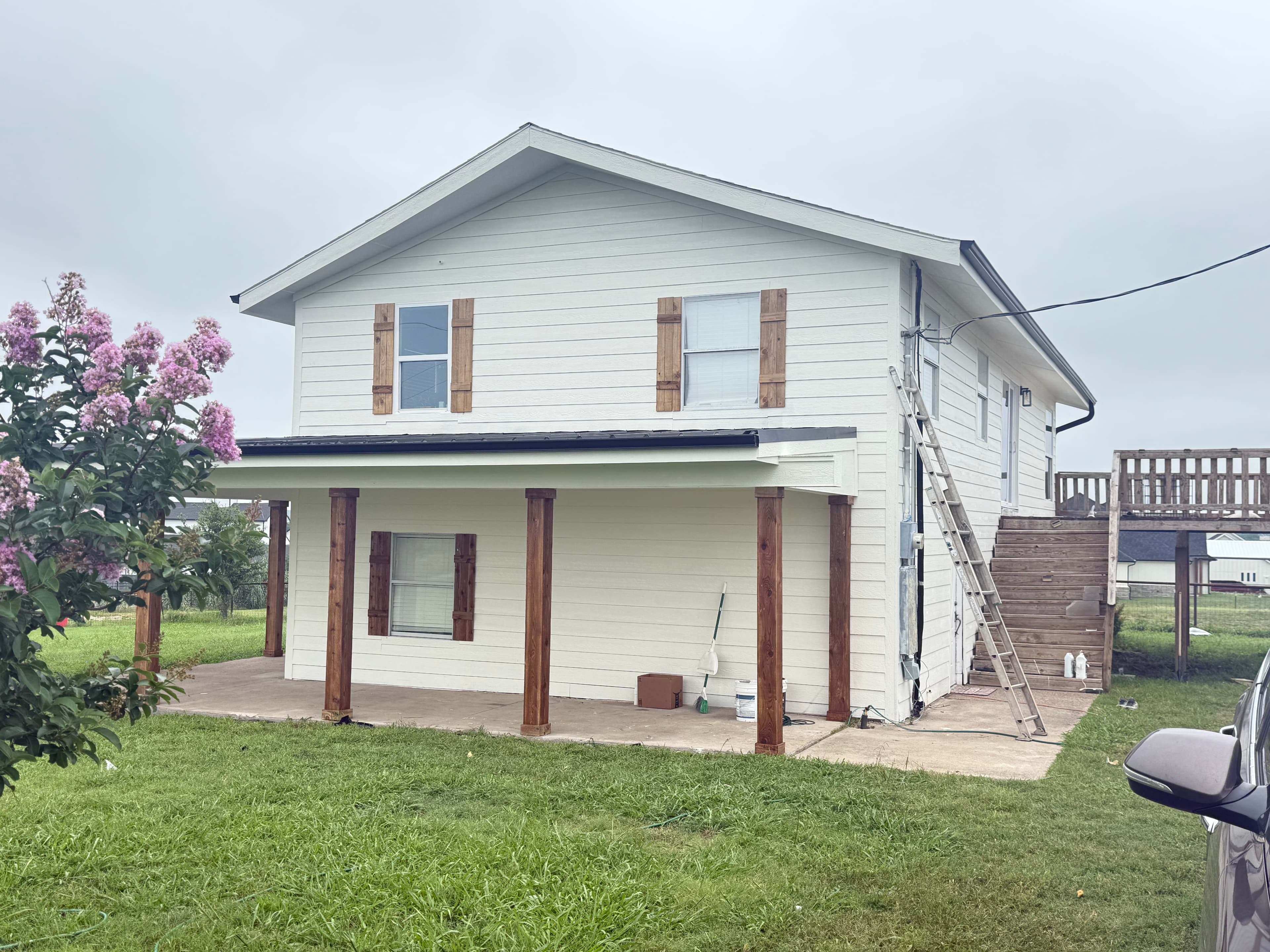 White house with brown shutters, wooden porch pillars, and a ladder leaning against the side.