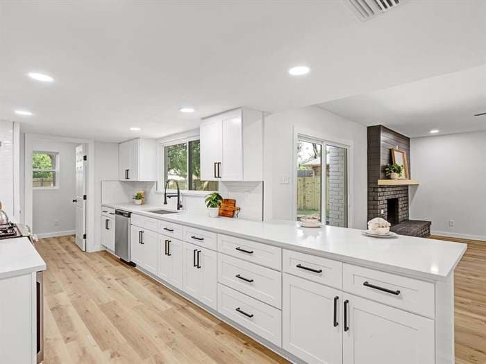 Modern white kitchen with black hardware, white countertops, light wood floors, and brick fireplace.