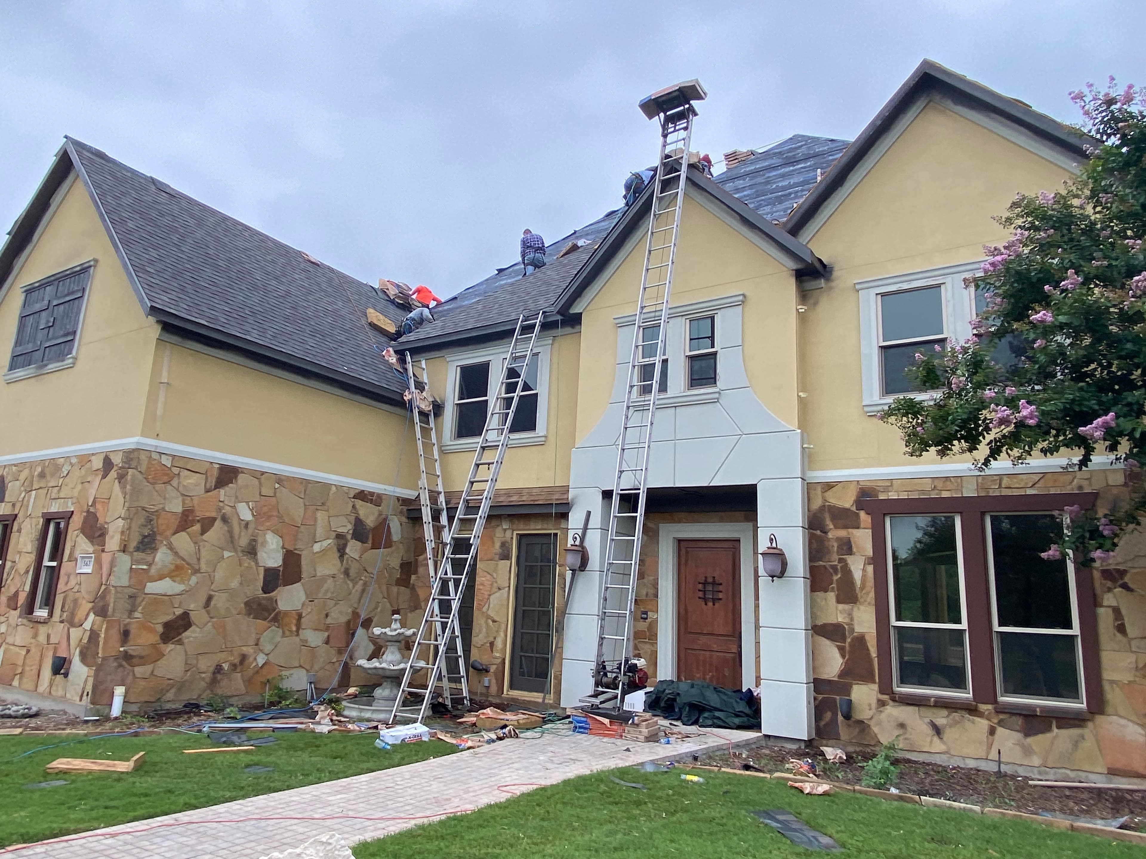 Roofers repair the roof of a large yellow and stone house using tall extension ladders.